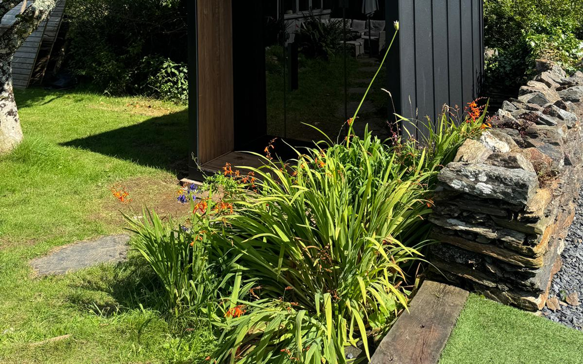 Summer garden approach towards a modern outdoor sauna threshold, used to inspect route questions.