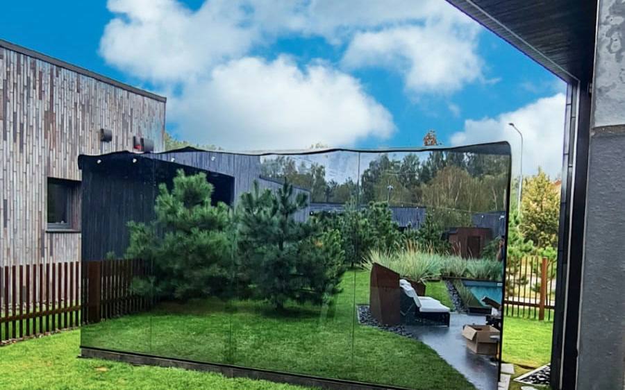 A mirrored panoramic cube sauna standing in a landscaped garden beside an existing building.