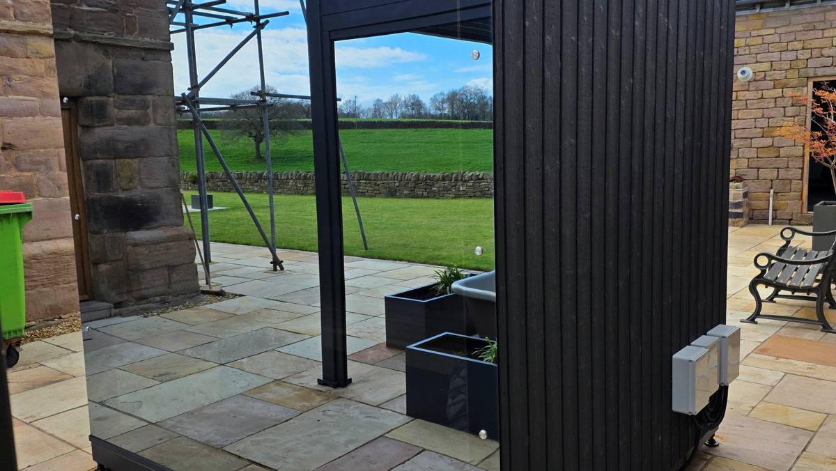 A dark glazed cube sauna standing on paving beside stone buildings and a garden.